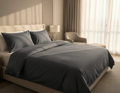 Luxe antraciet dekbedovertrek 240x220 lits-jumeaux, strak opgemaakt op een beige boxspring in een lichte moderne slaapkamer met hoge ramen, warme zonlichtinval, nachtkastjes en een stijlvolle fauteuil.